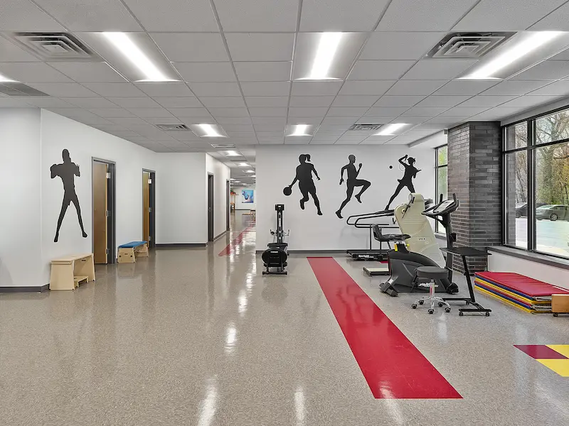 A large therapy gym with exercise equipment, including treadmills and stationary bikes. The space features bold red floor markings and black sports silhouettes painted on the walls. Bright windows line the room, allowing for ample natural light, and colorful therapy mats are stacked neatly in the corner.