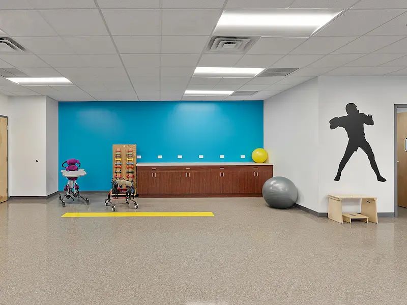 A therapy space with a turquoise accent wall and equipment including a therapy walker, an exercise ball, and a wooden balance beam. A storage cabinet runs along the wall beneath the bright blue section, and a black silhouette of a child playing sports decorates the adjacent white wall.