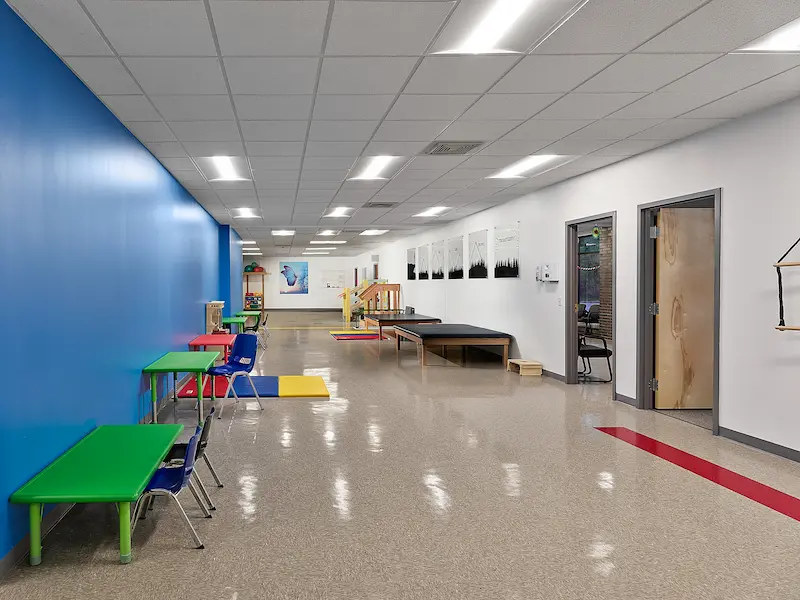 A spacious therapy area with a long hallway, vibrant blue walls, and green and red kids' tables with chairs. The floor features colorful mats, and there is a small wooden climbing ramp at the far end of the room. The bright lighting and open layout are designed for physical therapy and child activities.