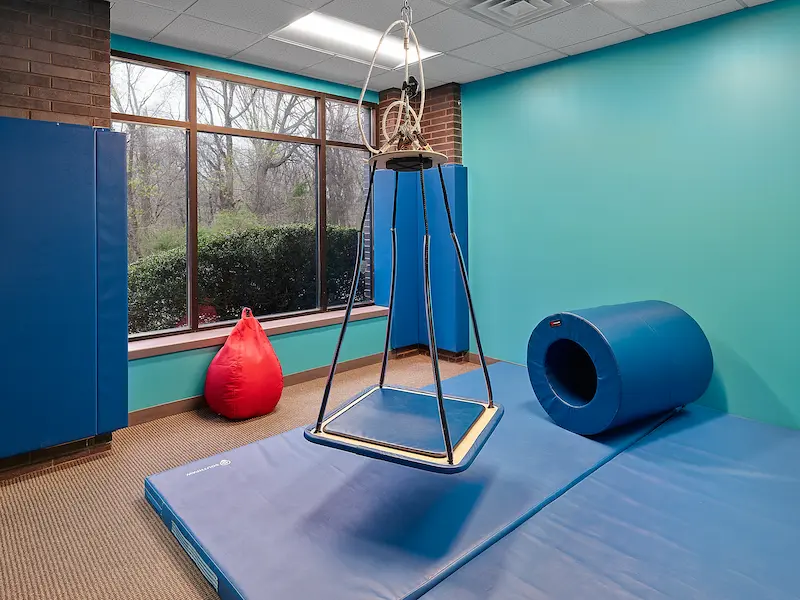 A sensory therapy room featuring a hanging platform swing suspended from the ceiling. The room has padded blue mats covering the floor and walls for safety. A large blue foam cylinder lies on the floor, and a red beanbag sits near a window with trees visible outside.