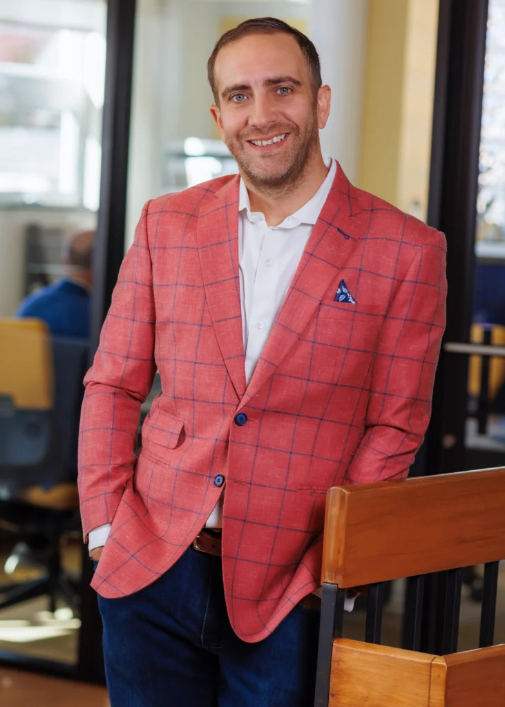 A man in a red checkered blazer and white shirt, leaning casually on a wooden handrail in an office. He is smiling warmly, with one hand resting in his pocket.