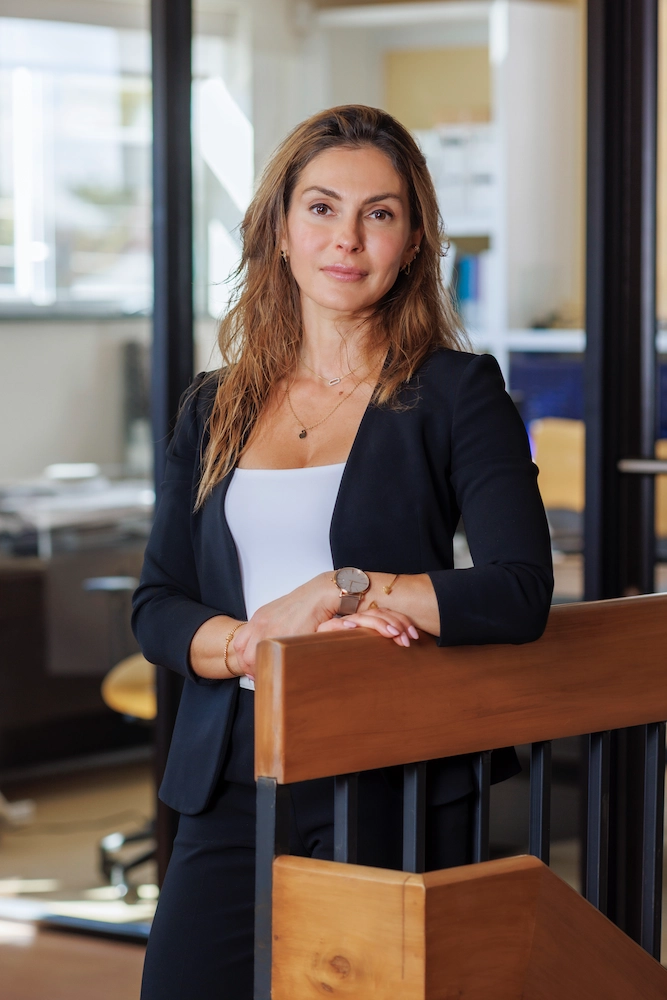 A woman in a black blazer and white top, leaning against a wooden handrail in an office environment. She has a calm expression, wearing subtle jewelry, and her arms are crossed lightly.
