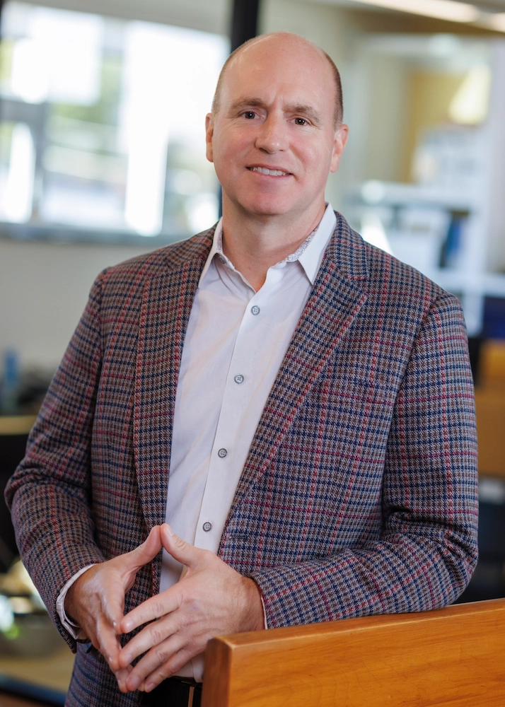 A man in a plaid blazer with a white shirt, standing confidently near a wooden handrail in an office setting. He has a light smile and his hands are clasped together.